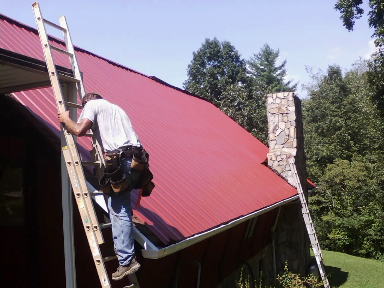 Roofer on a ladder beside a red metal roof and stone chimney — Soffit & Fascia Repair by Roofing Brothers in Lisbon Falls